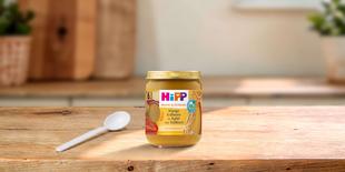 HiPP Bio-Gläschen Mango Erdbeere in Apfel mit Vollkorn auf Holztisch, daneben ein Löffel in heller Küchenumgebung