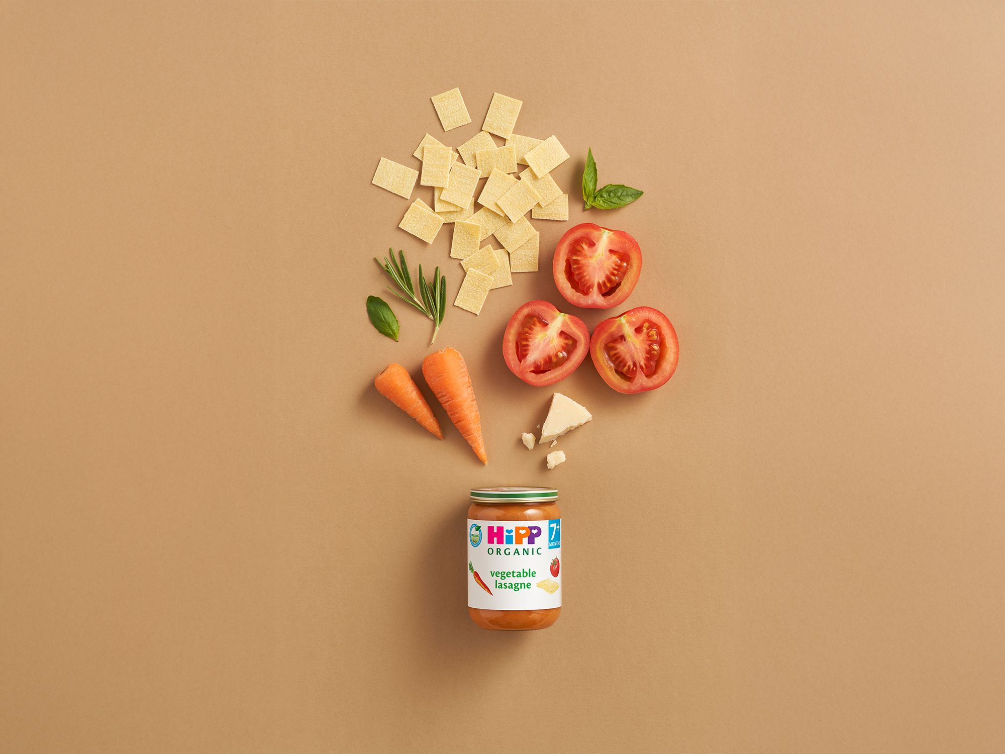 HiPP Organic Vegetable Lasagne jar with pasta, tomatoes, carrots, herbs and cheese arranged above on a neutral background