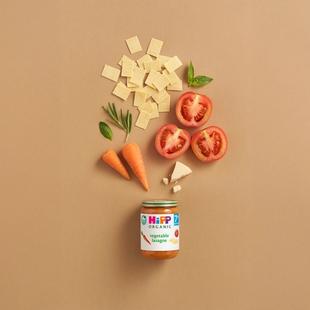 HiPP Organic Vegetable Lasagne jar with pasta, tomatoes, carrots, herbs and cheese arranged above on a neutral background