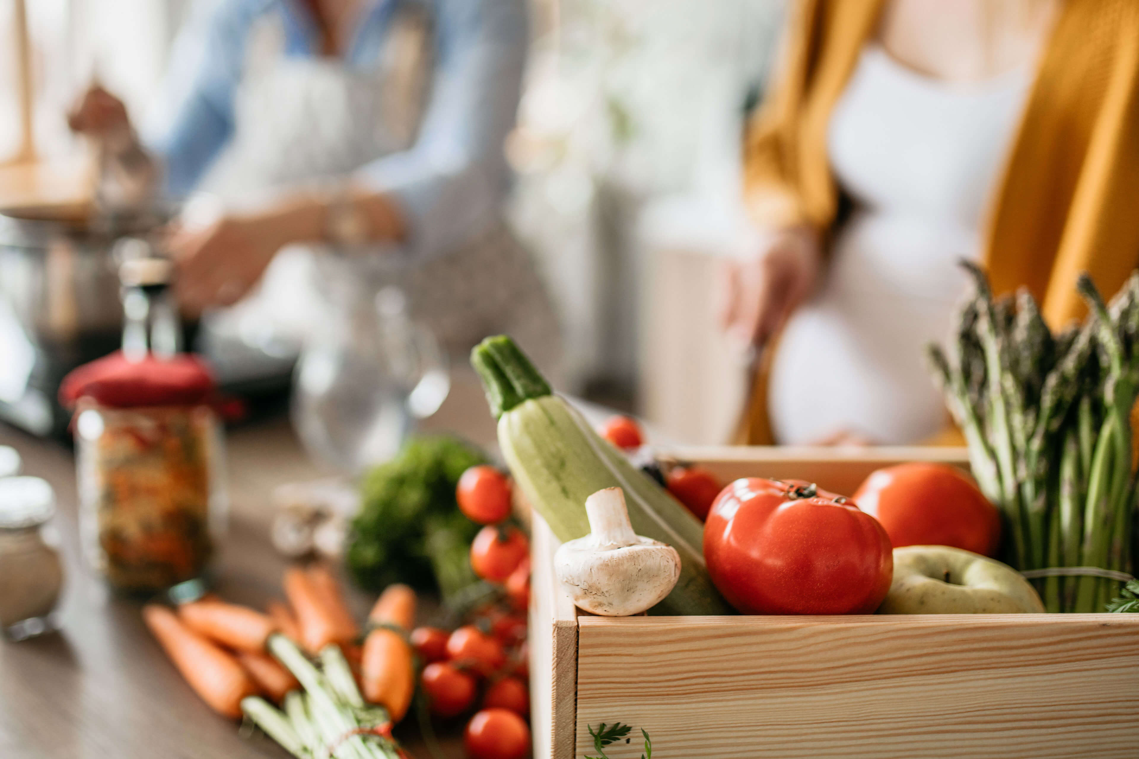 Eine Holzkiste, gefüllt mit frischem Gemüse, darunter Tomaten, Spargel und Knoblauch, steht auf einer Küchenzeile.