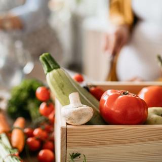 Eine Holzkiste, gefüllt mit frischem Gemüse, darunter Tomaten, Spargel und Knoblauch, steht auf einer Küchenzeile.
