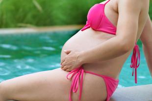 Eine Frau in einem rosafarbenen Bikini, die sanft ihren schwangeren Bauch an einem Pool berührt.