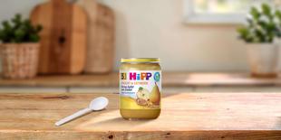 HiPP Bio Frucht & Getreide Glas Birne-Apfel mit Dinkel auf Holztisch mit Babylöffel in heller Küche