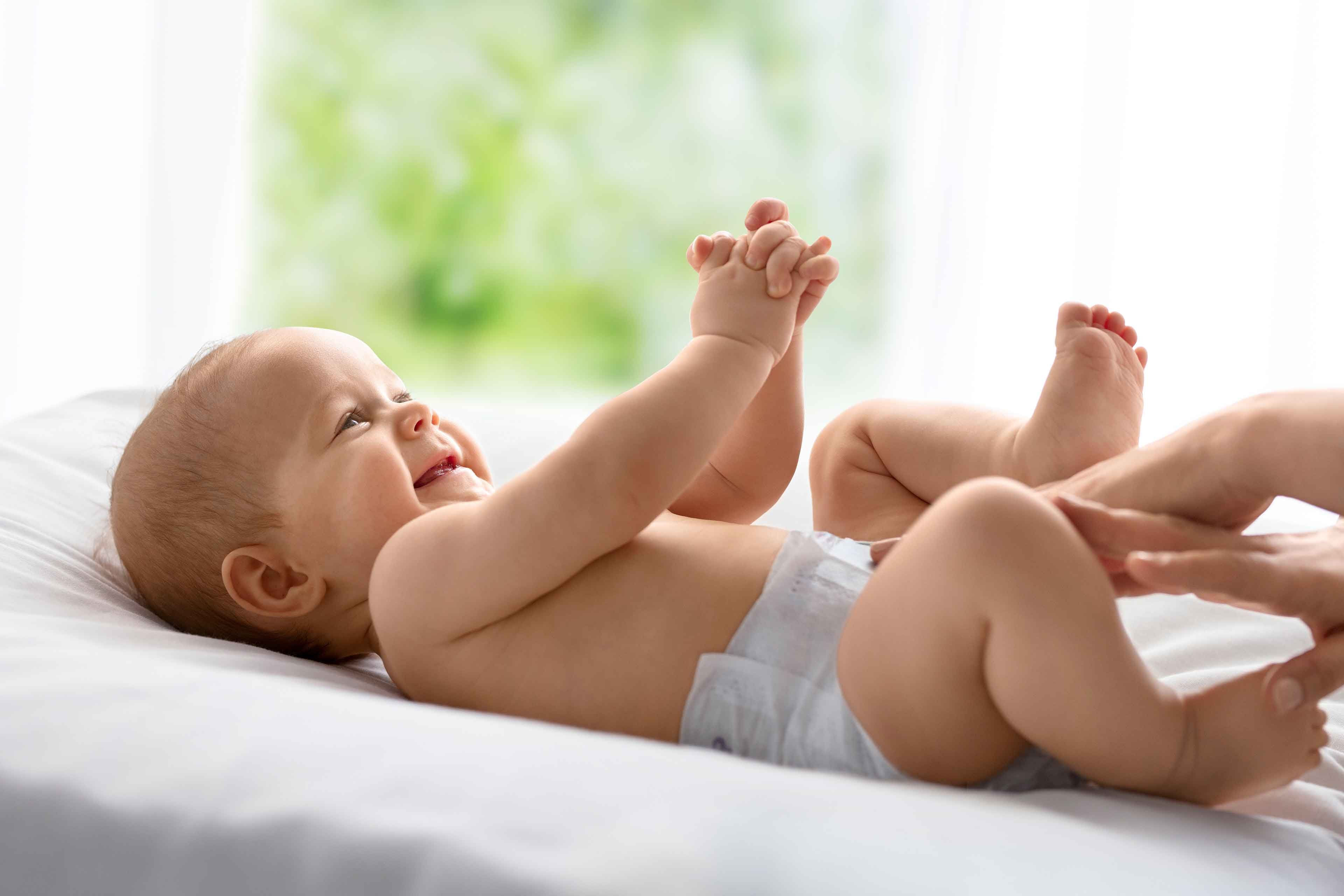 Ein fröhliches Baby, das auf einem Bett liegt und beide Hände und Füße ausstreckt, während die Hand einer Person sanft den Fuß des Babys berührt.