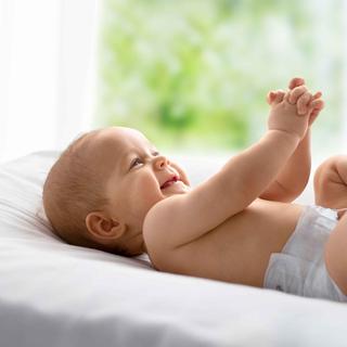 Ein fröhliches Baby, das auf einem Bett liegt und beide Hände und Füße ausstreckt, während die Hand einer Person sanft den Fuß des Babys berührt.