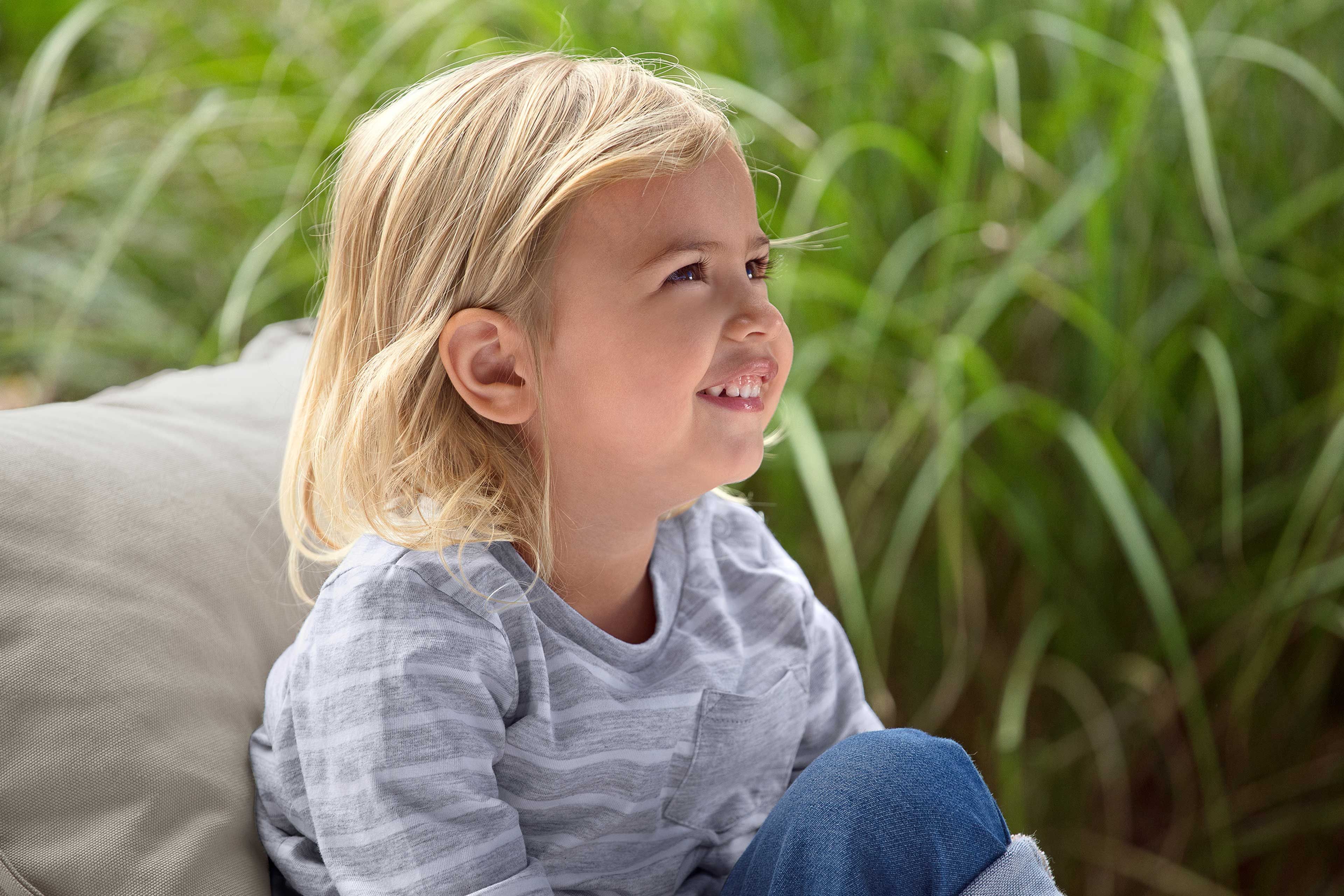 Ein kleines Kind mit blondem Haar, das ein gestreiftes Hemd und eine blaue Jeans trägt, sitzt im Freien inmitten von hohem grünen Gras.