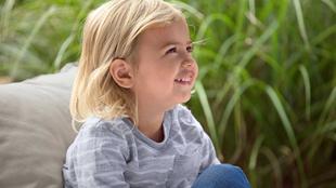 Ein kleines Kind mit blondem Haar, das ein gestreiftes Hemd und eine blaue Jeans trägt, sitzt im Freien inmitten von hohem grünen Gras.