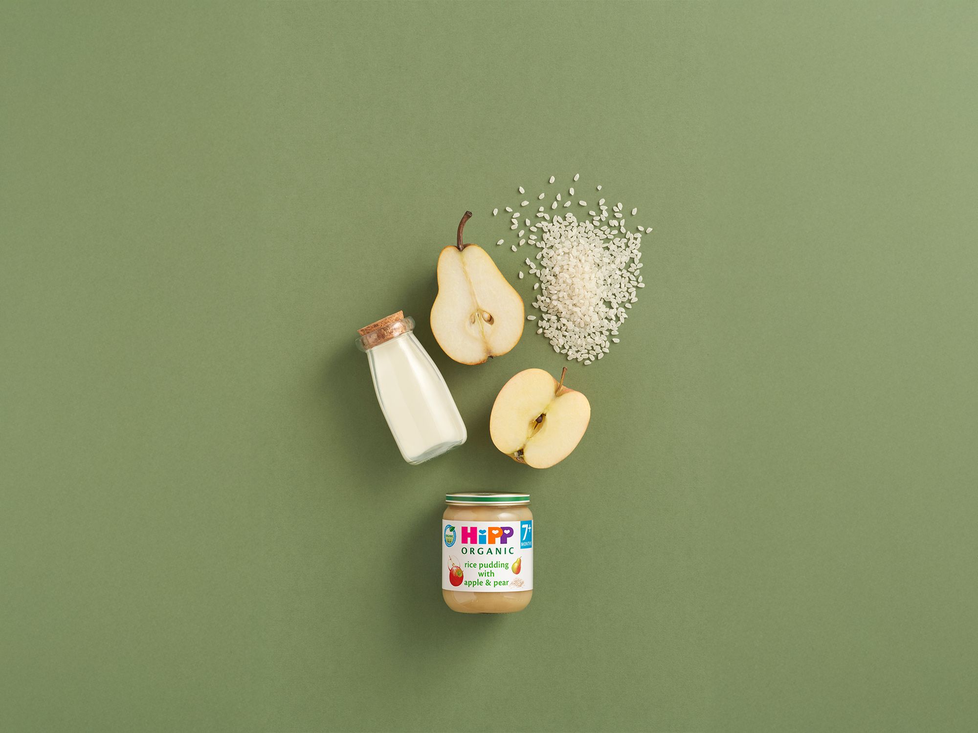 HiPP Organic Rice Pudding with Apple & Pear jar styled with milk, pear halves, apple slices and rice on a green background