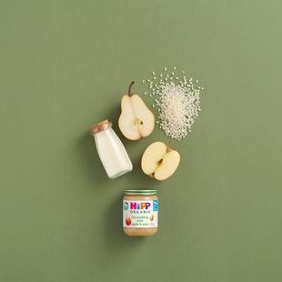 HiPP Organic Rice Pudding with Apple & Pear jar styled with milk, pear halves, apple slices and rice on a green background
