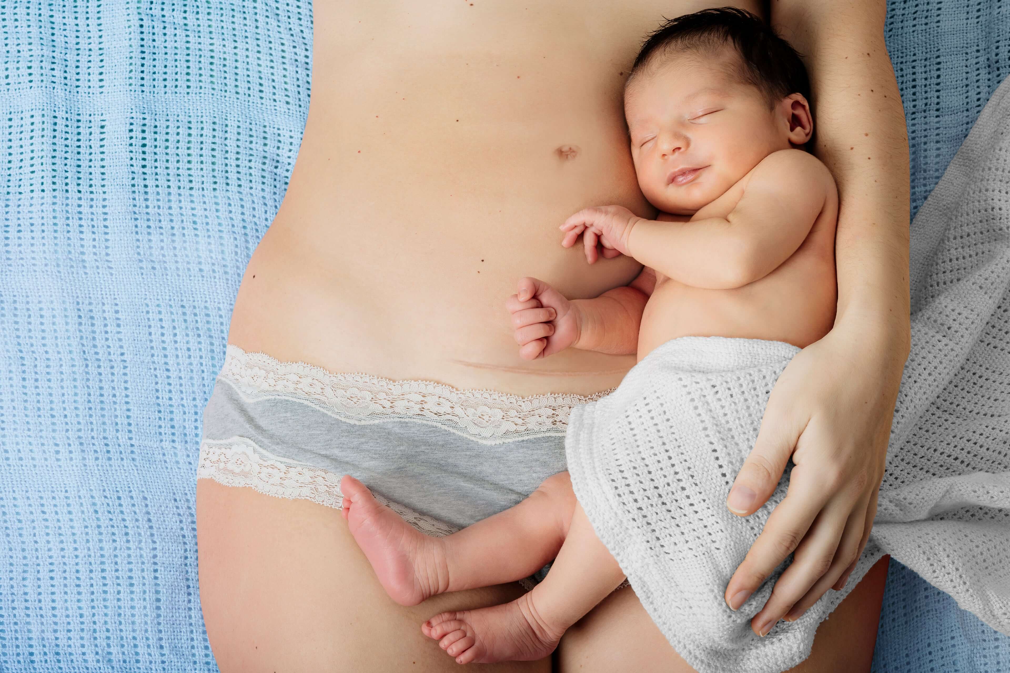 Eine Mutter hält ihr neugeborenes Baby, das in ein weißes Tuch gewickelt ist, während die Mutter hellblaue Unterwäsche mit Spitzenverzierung trägt.