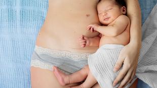 Eine Mutter hält ihr neugeborenes Baby, das in ein weißes Tuch gewickelt ist, während die Mutter hellblaue Unterwäsche mit Spitzenverzierung trägt.