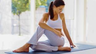 Eine Frau in einem weißen Sport-BH und einer grauen Hose sitzt auf einer Yogamatte, in entspannter Haltung und mit einem gelassenen Gesichtsausdruck.