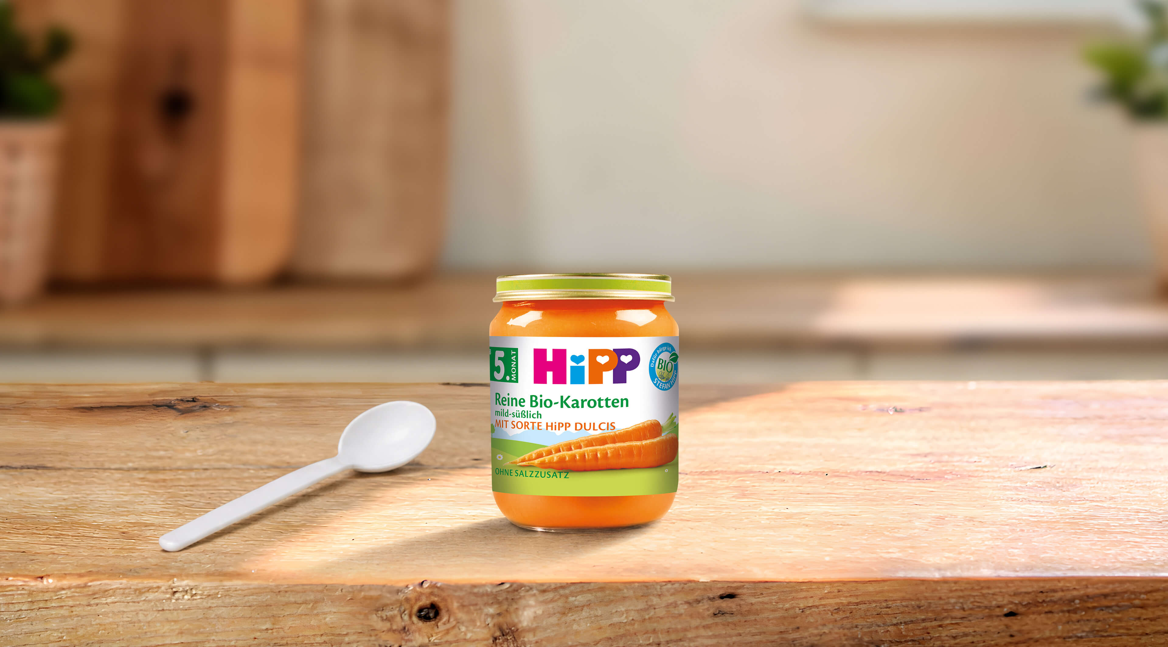 HiPP Reine Bio-Karotten Babygläschen ab dem 5. Monat mit Löffel auf einem Holztisch in heller Küchenumgebung
