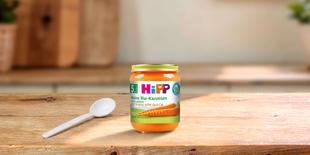 HiPP Reine Bio-Karotten Babygläschen ab dem 5. Monat mit Löffel auf einem Holztisch in heller Küchenumgebung