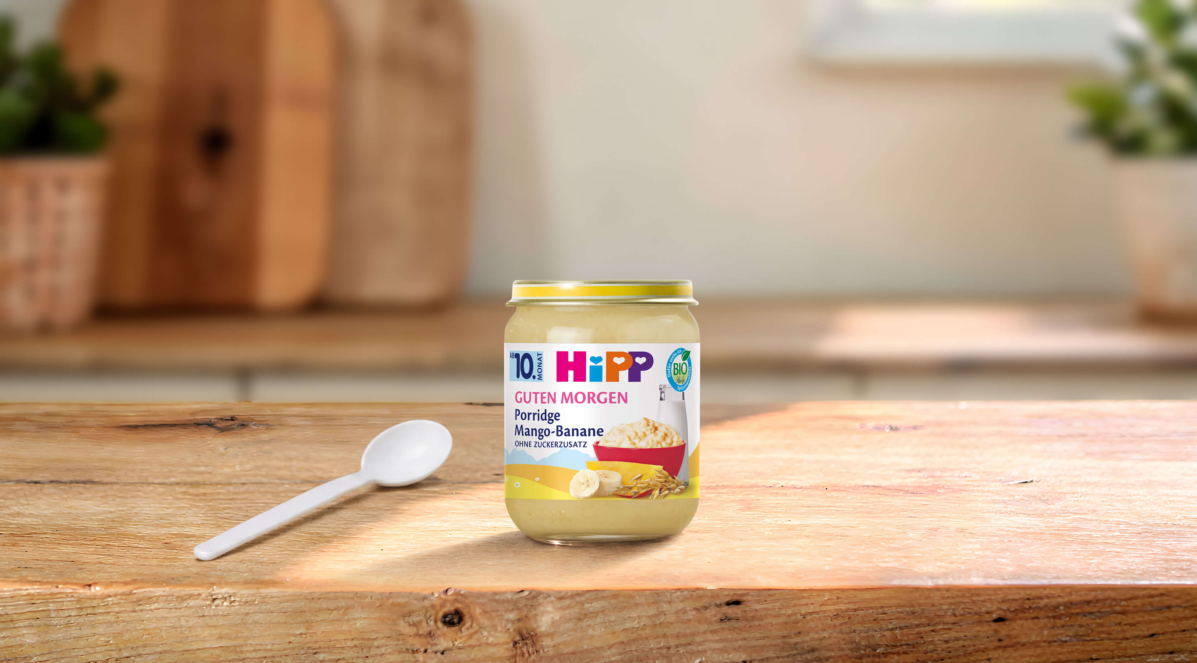 HiPP Guten Morgen Bio-Porridge Mango-Banane im Glas mit Löffel auf Holztisch in heller Küche