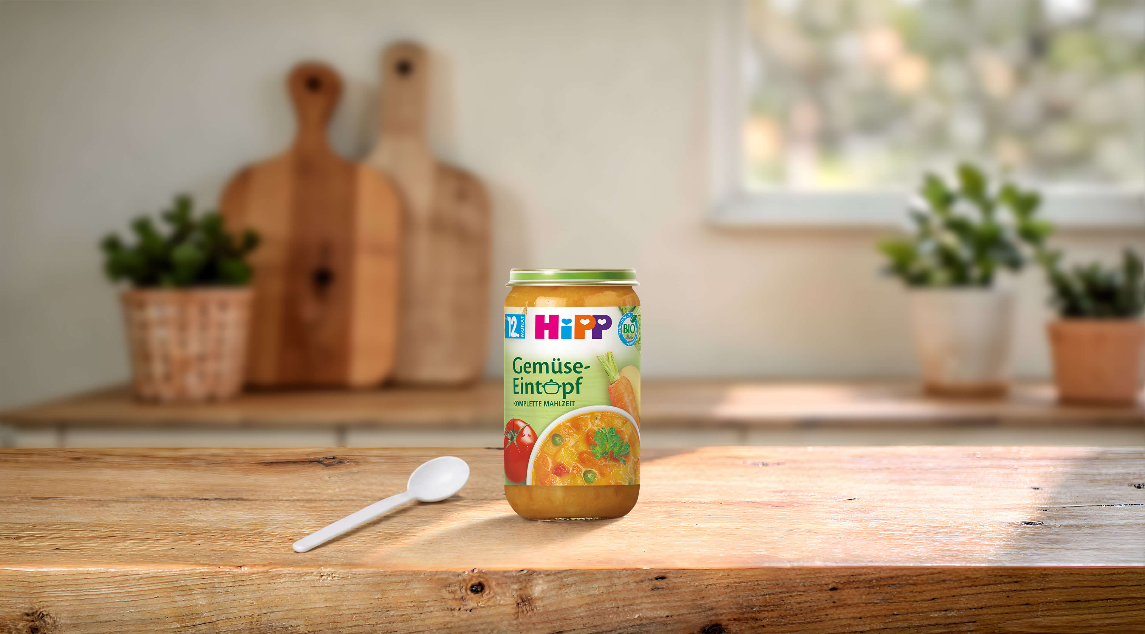 HiPP Bio Gemüse-Eintopf im 250g-Glas auf Holztisch mit Löffel in heller, gemütlicher Küche arrangiert