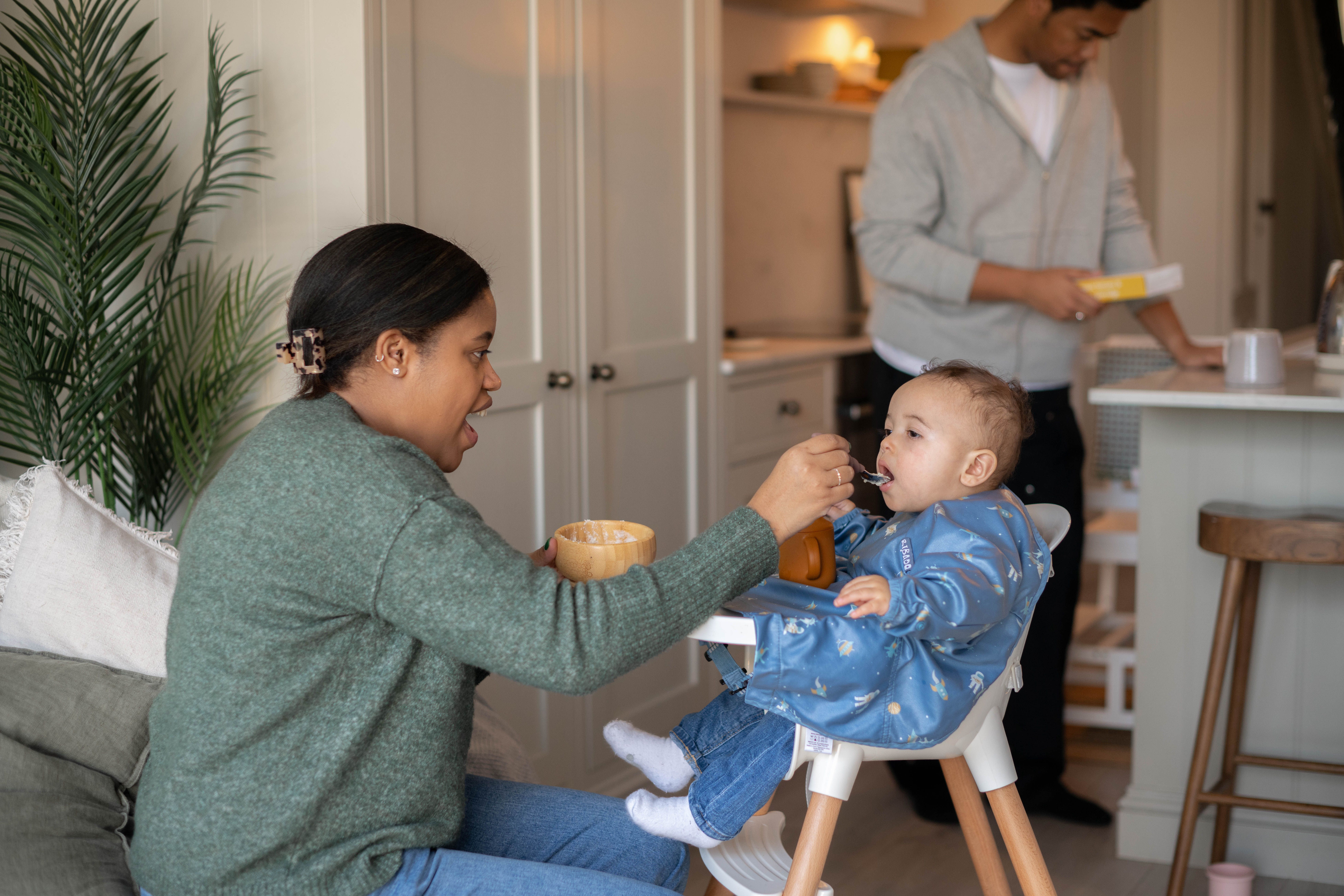 Parent feeding a baby in a highchair during weaning, with another parent preparing food in a cosy kitchen