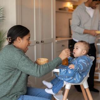Parent feeding a baby in a highchair during weaning, with another parent preparing food in a cosy kitchen