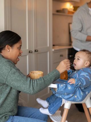 Parent feeding a baby in a highchair during weaning, with another parent preparing food in a cosy kitchen