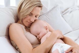 Mutter liegt entspannt im Bett und hält ihr schlafendes Baby im Arm, beide kuscheln eng in ruhiger, heller Umgebung
