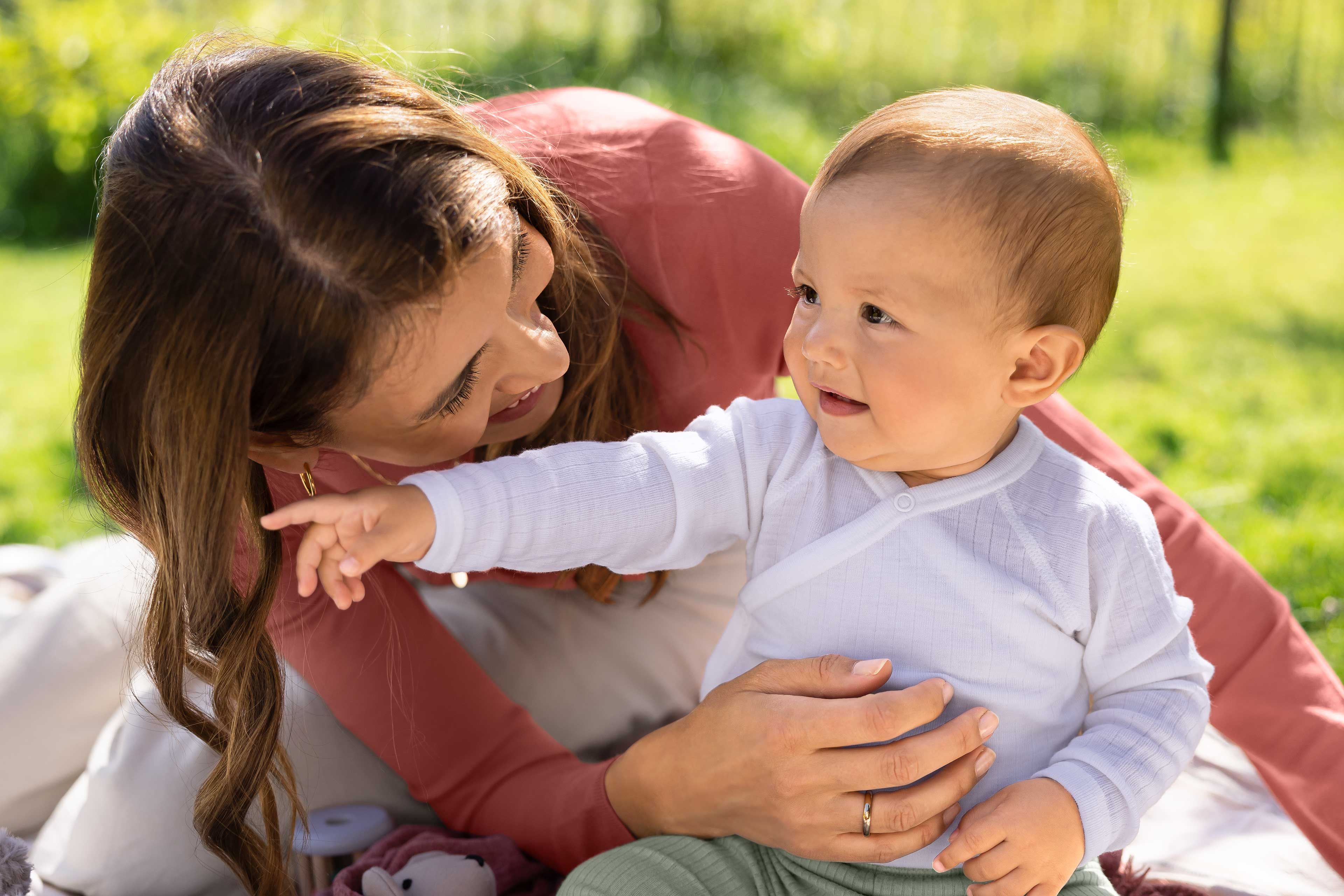 Mutter hält ihr lachendes Baby im Freien, beide schauen sich liebevoll an auf einer Picknickdecke