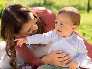 Mutter hält ihr lachendes Baby im Freien, beide schauen sich liebevoll an auf einer Picknickdecke