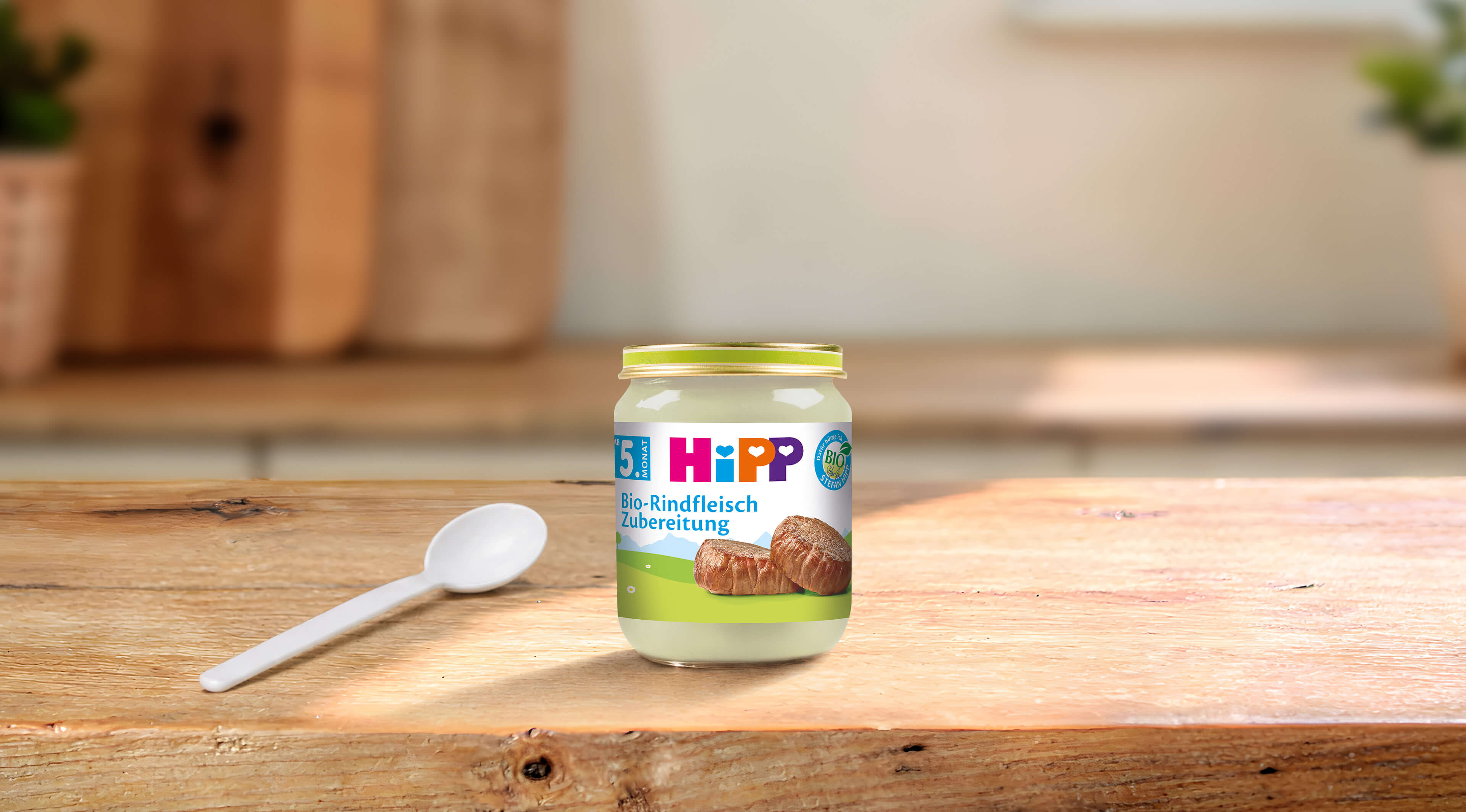 HiPP Bio-Rindfleisch Zubereitung im 125g-Glas mit Löffel auf einem Holztisch in heller Küchenumgebung