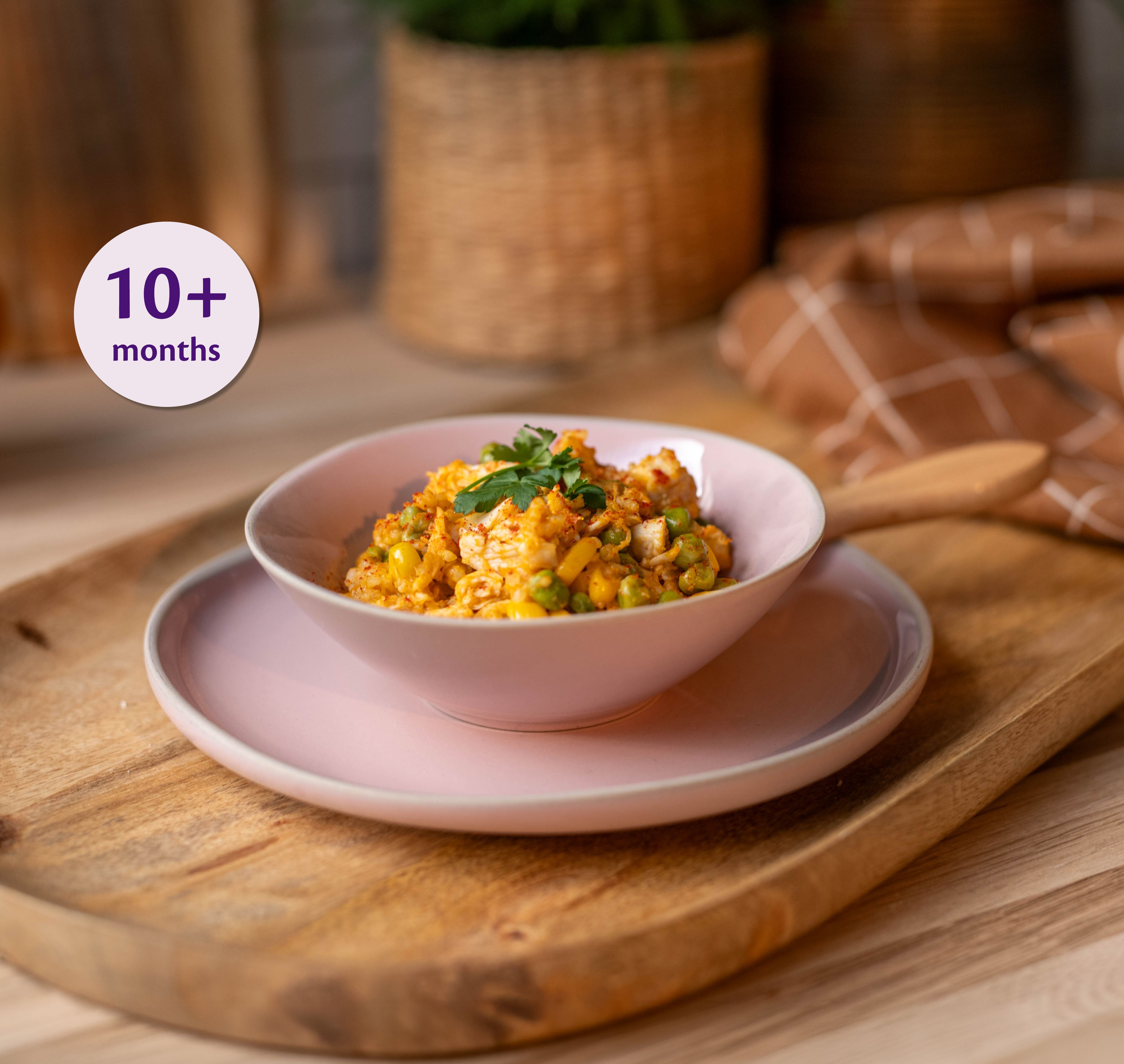 Small bamboo bowl of soft chicken and veg paella for babies, with peas and rice, beside a baby spoon on a marble surface