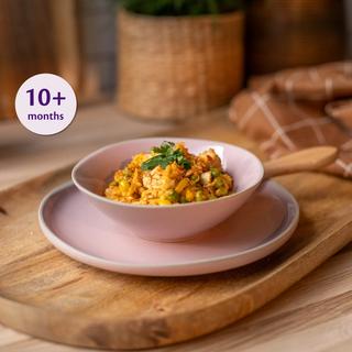 Small bamboo bowl of soft chicken and veg paella for babies, with peas and rice, beside a baby spoon on a marble surface