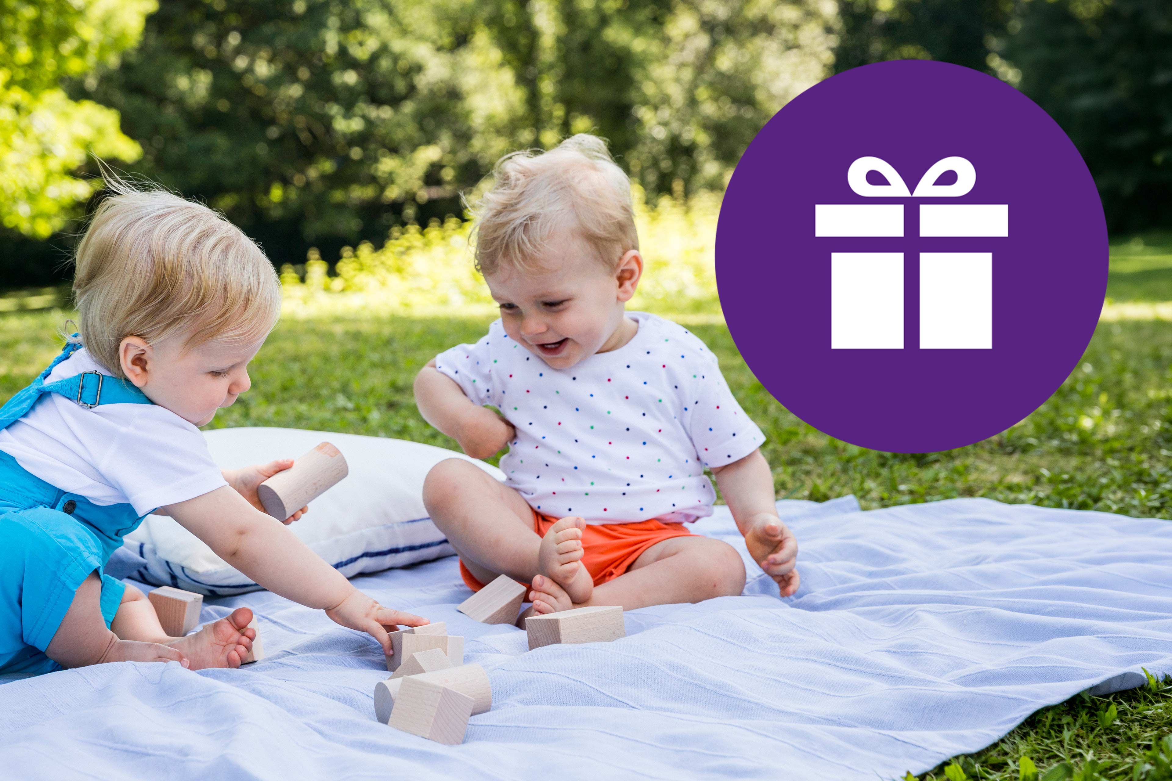 Zwei Kleinkinder sitzen auf einer Decke im Freien und spielen mit Holzbausteinen, daneben ein lila Kreis mit weißem Geschenk-Symbol