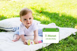 Ein Baby im Alter von 7 Monaten, das mit einem bunten Spielzeug auf einer Decke im Freien spielt.