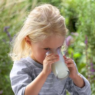 Kleines blondes Kind trinkt draußen ein Glas Milch, umgeben von grünem Garten im Hintergrund