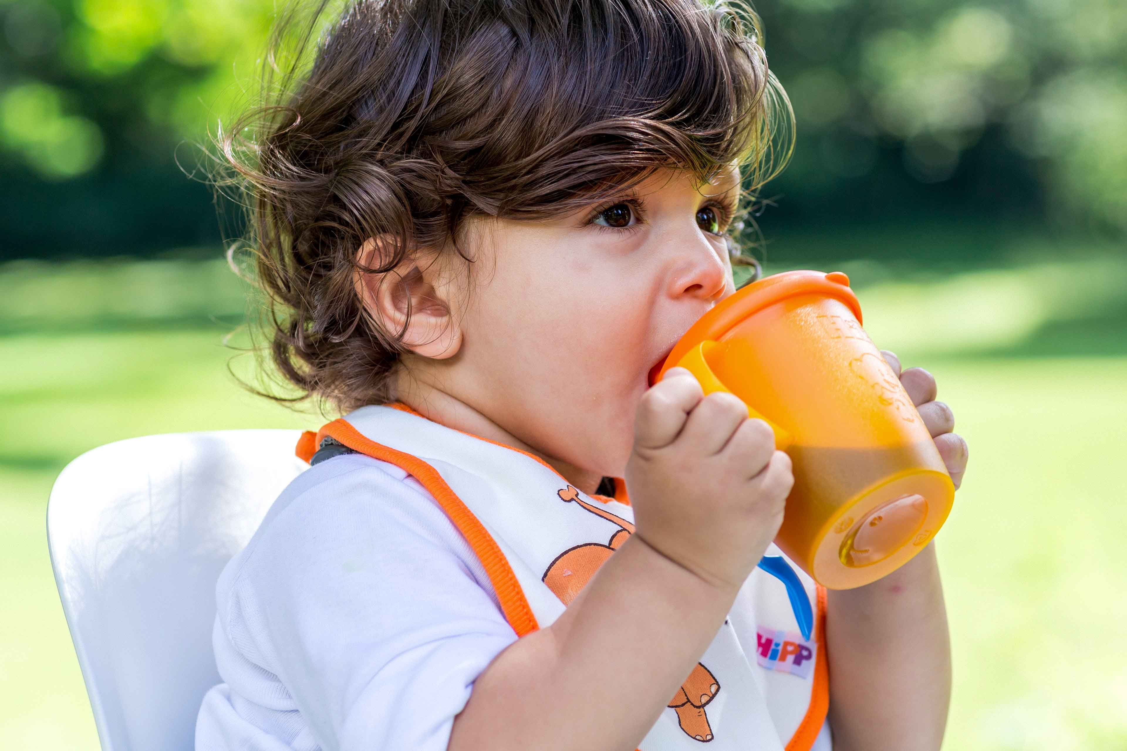 Ein kleines Kind, vermutlich ein Kleinkind, nippt im Freien an einer orangefarbenen Schnabeltasse.