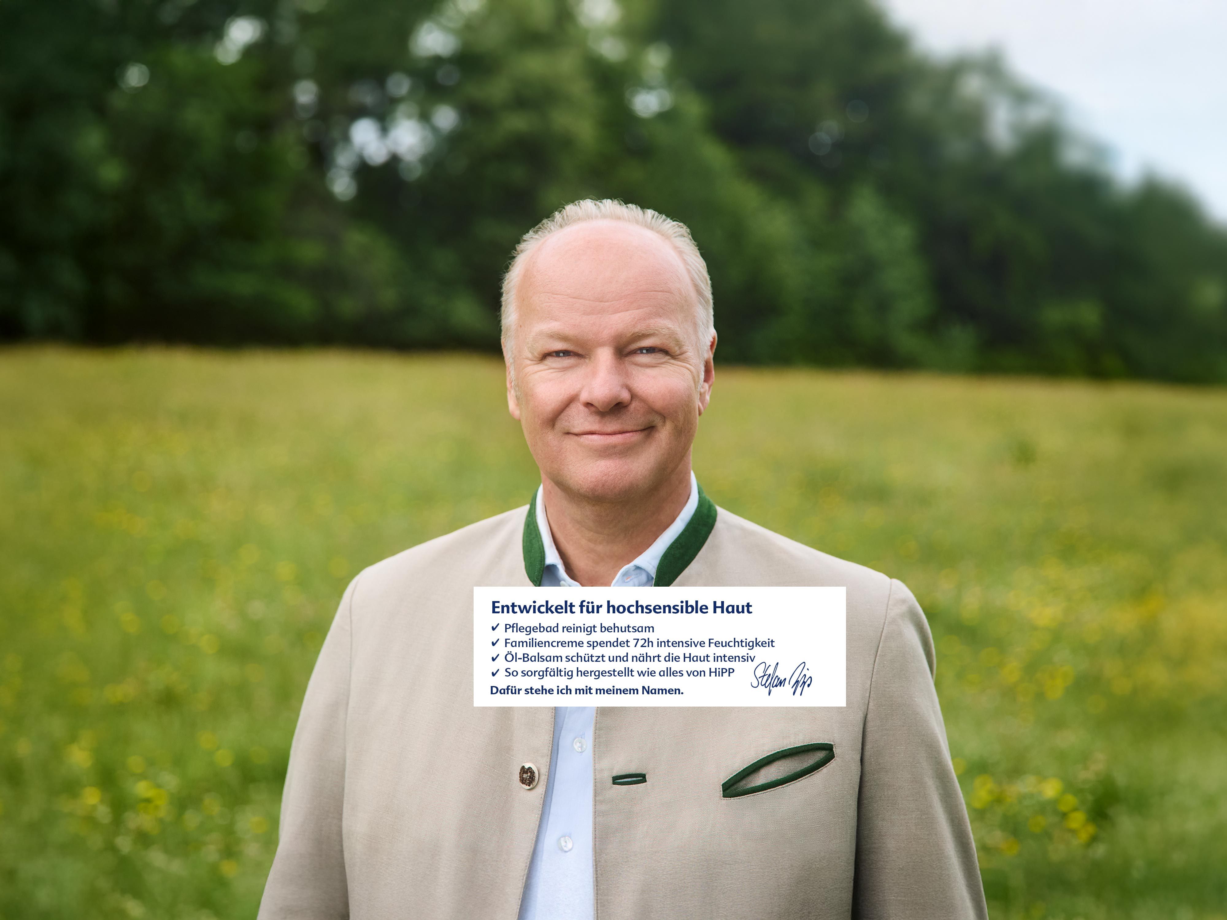 Mann in traditioneller Trachtenjacke steht auf einer Wiese vor Bäumen und lächelt