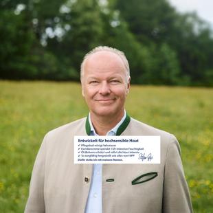 Mann in traditioneller Trachtenjacke steht auf einer Wiese vor Bäumen und lächelt