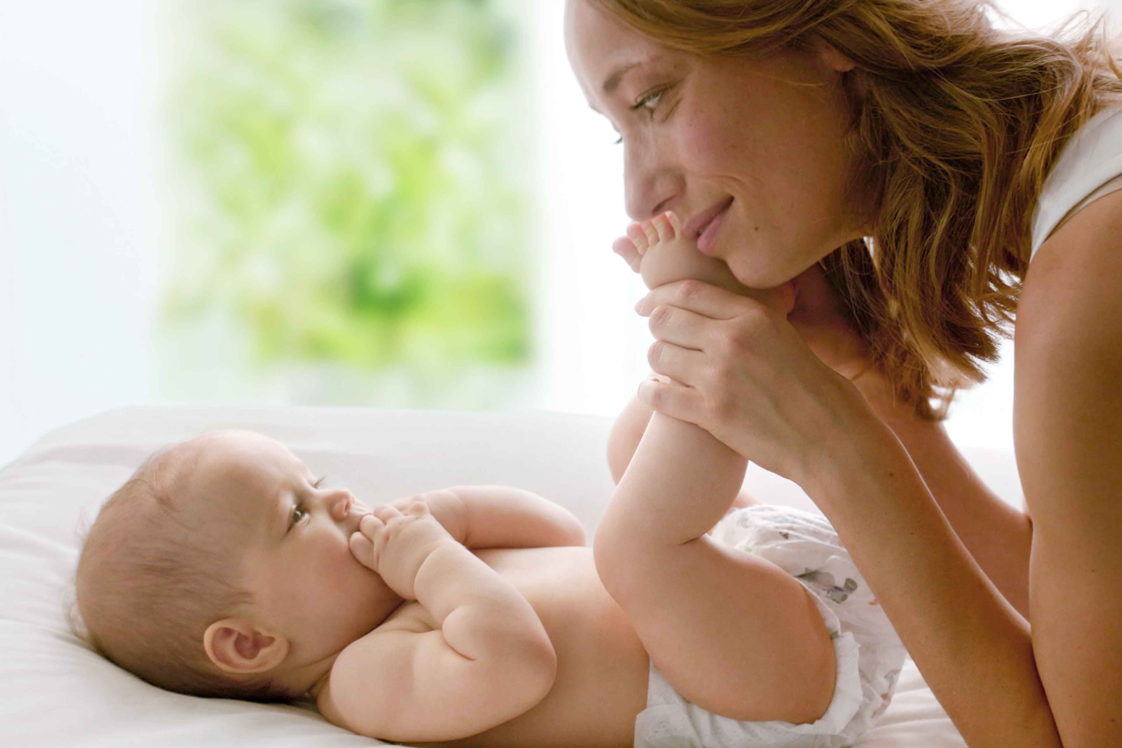 Ein Baby, das auf einer weißen Fläche liegt und sich an seinen eigenen Füßen festhält, während die Hand einer Person sanft sein Bein berührt.