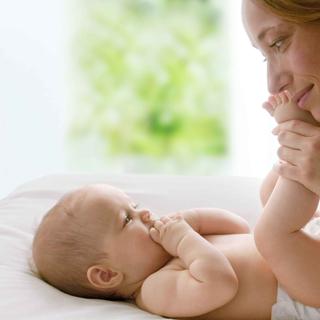 Ein Baby, das auf einer weißen Fläche liegt und sich an seinen eigenen Füßen festhält, während die Hand einer Person sanft sein Bein berührt.