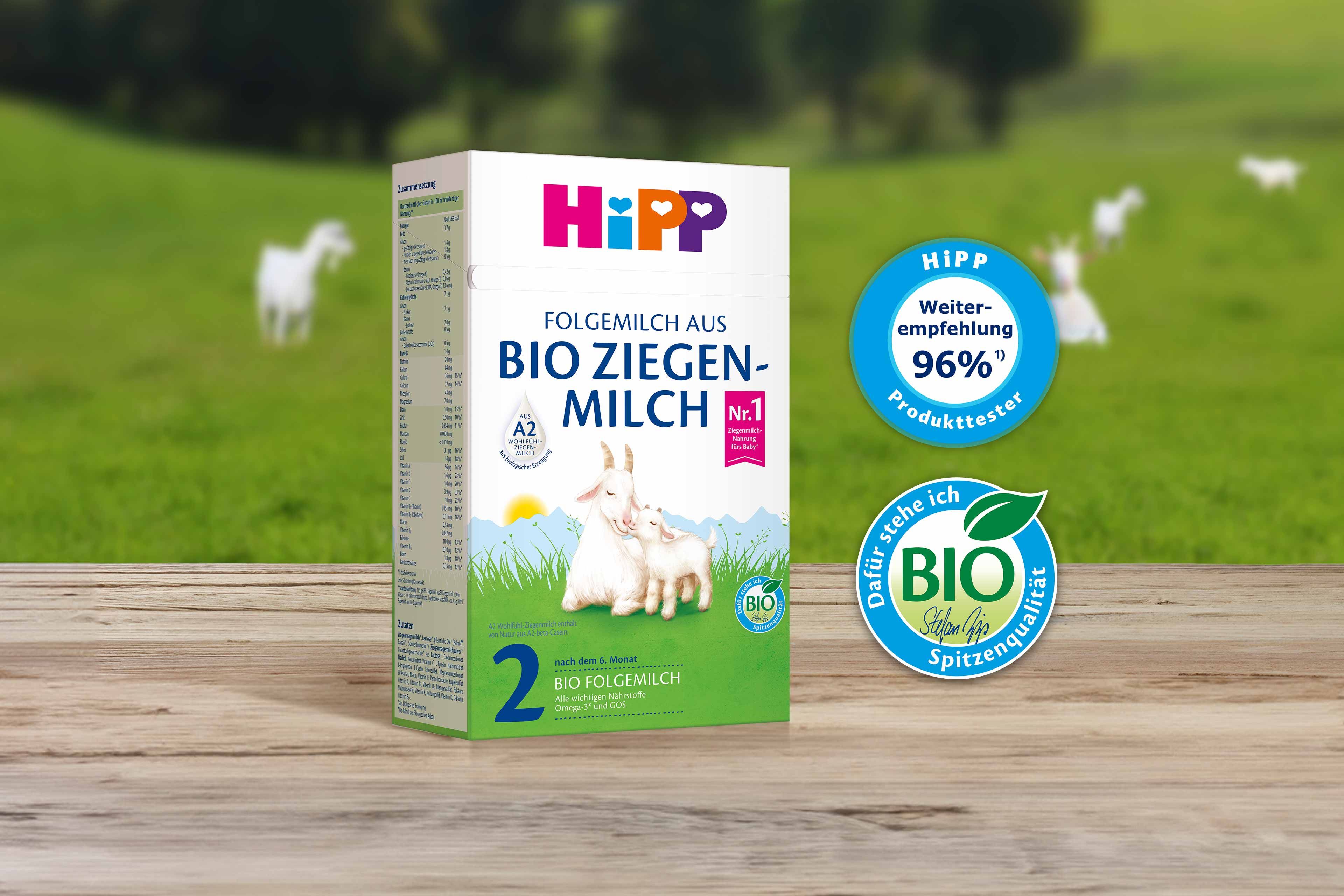 HiPP 2 Folgemilch aus Bio Ziegenmilch auf Holztisch, daneben Bio- und Produkttester-Siegel, im Hintergrund grüne Weide mit Ziegen.