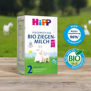 HiPP 2 Folgemilch aus Bio Ziegenmilch auf Holztisch, daneben Bio- und Produkttester-Siegel, im Hintergrund grüne Weide mit Ziegen.
