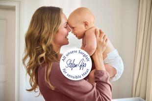 Lächelnde Mutter hält ihr Baby hautnah im Arm, beide Stirn an Stirn, mit Siegel „All unsere Sorgfalt für sensible Haut“ im Vordergrund