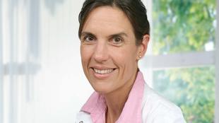 Eine Frau mit strahlendem Lächeln, die ein weißes Hemd mit rosa Kragen trägt, vor einem hellen Hintergrund.