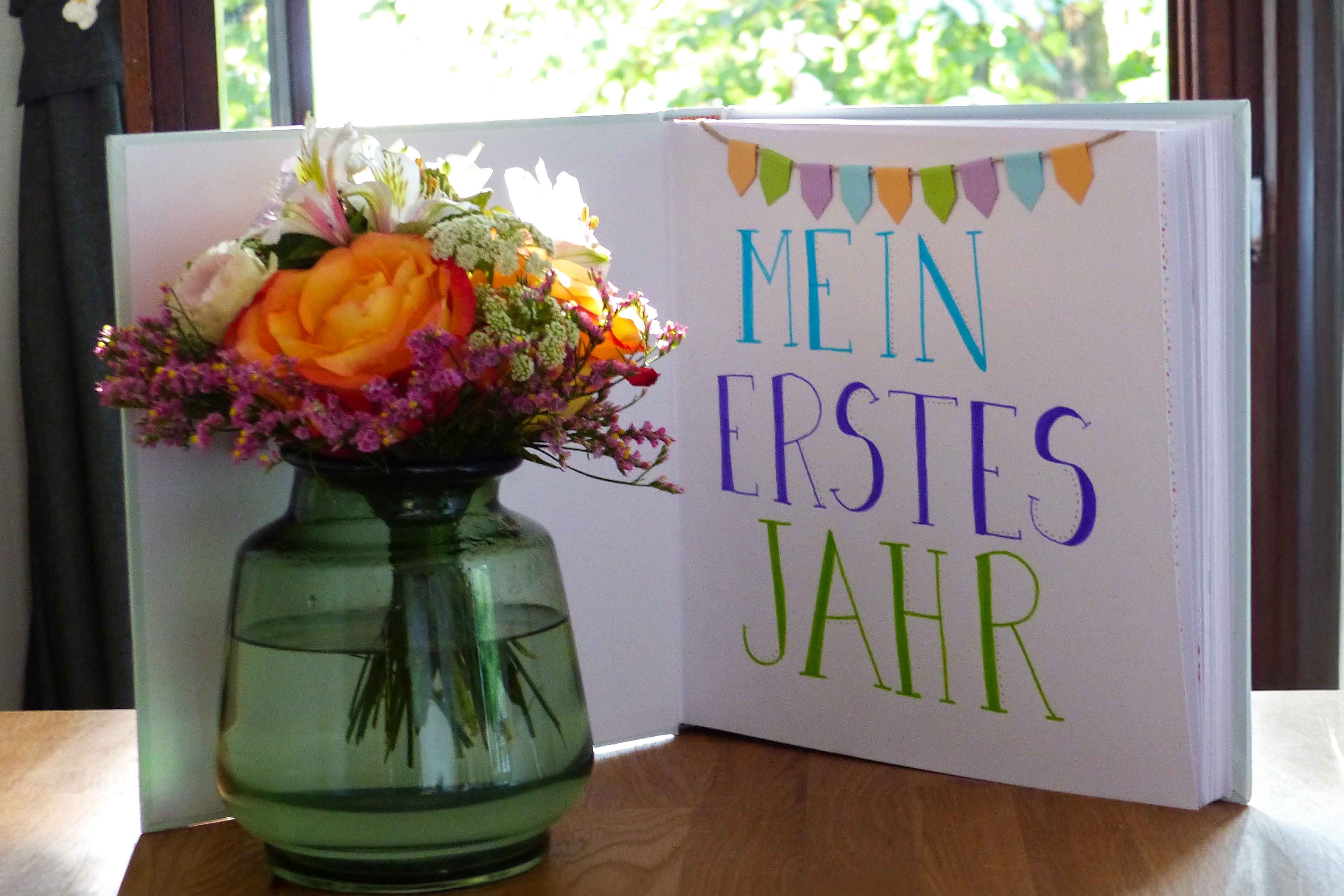 Ein Strauß leuchtender Blumen neben einem Buch mit dem Titel "Mein erstes Jahr" in deutscher Sprache, begleitet von bunten Bannern.