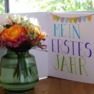 Ein Strauß leuchtender Blumen neben einem Buch mit dem Titel "Mein erstes Jahr" in deutscher Sprache, begleitet von bunten Bannern.