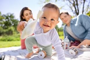 Lachendes Baby krabbelt auf einer Picknickdecke im Grünen, während die Eltern dahinter liebevoll mit ihm spielen.