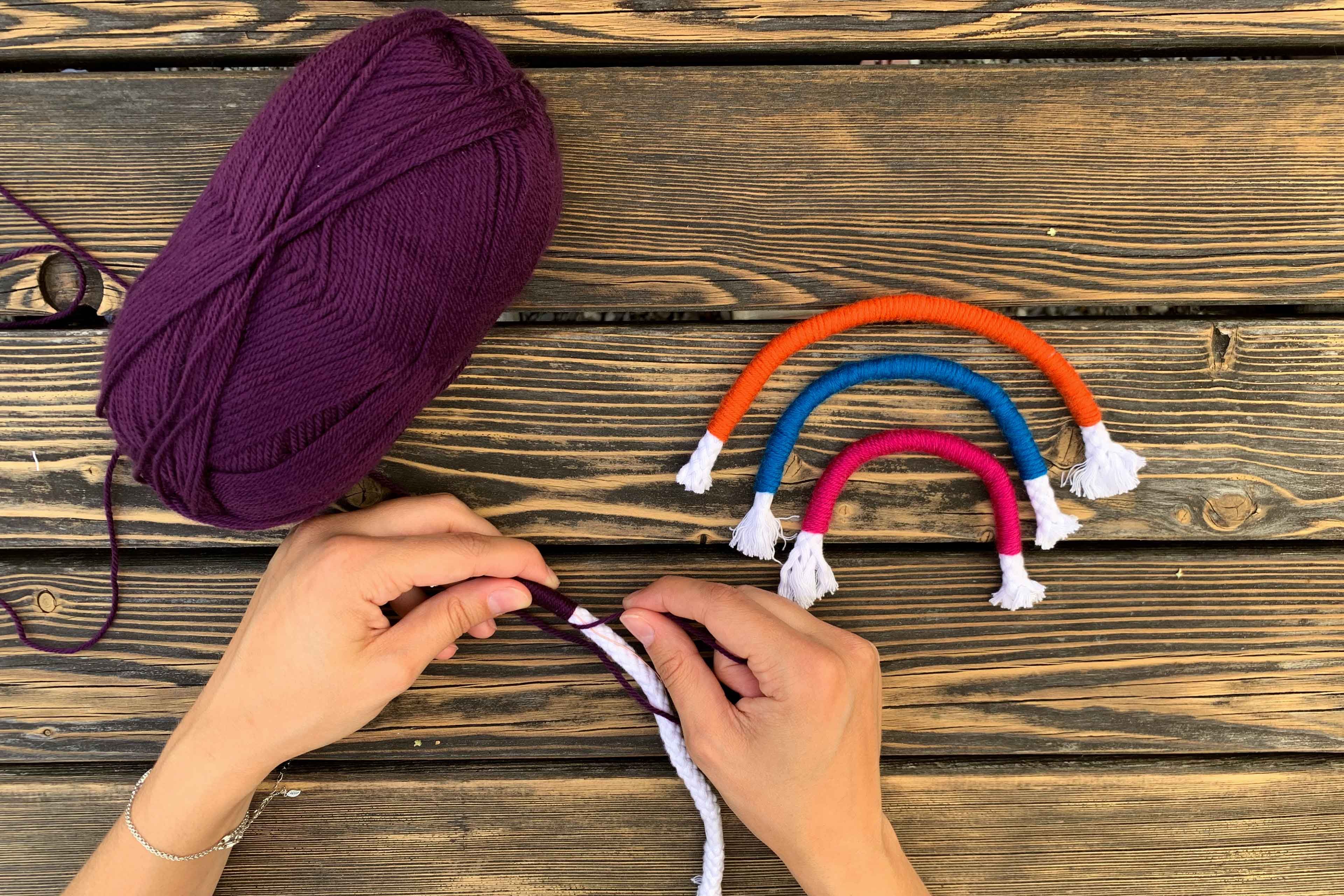 Die Hände einer Person, die mit einem violetten Garn arbeiten und ein gestricktes Objekt schaffen, vor dem Hintergrund einer hölzernen Oberfläche und einiger bunter gestrickter Ringe.
