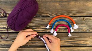 Die Hände einer Person, die mit einem violetten Garn arbeiten und ein gestricktes Objekt schaffen, vor dem Hintergrund einer hölzernen Oberfläche und einiger bunter gestrickter Ringe.