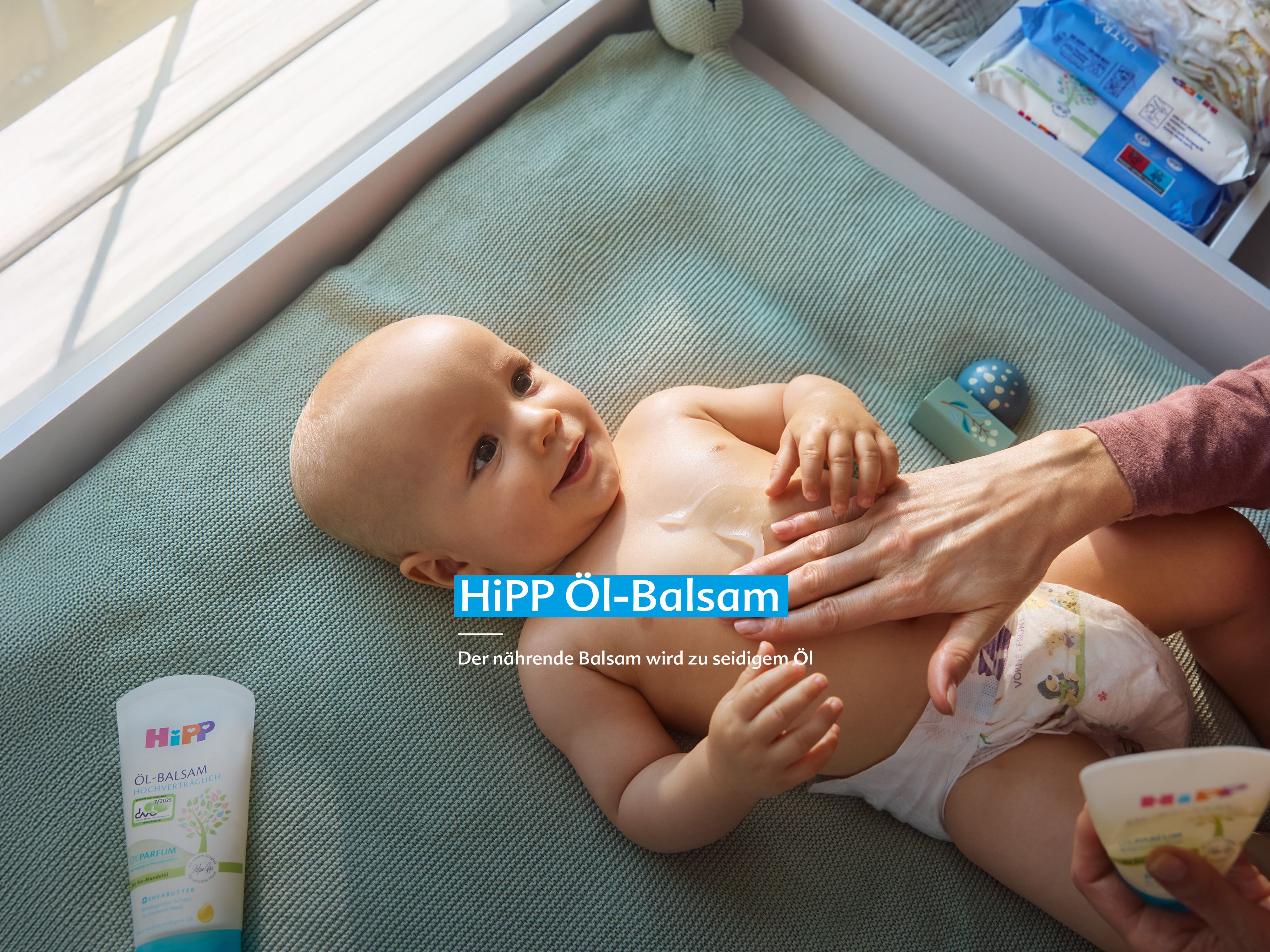 Lächelndes Baby auf Wickelauflage, dessen Bauch mit HiPP HOCHVERTRÄGLICH Öl-Balsam sanft eingecremt wird