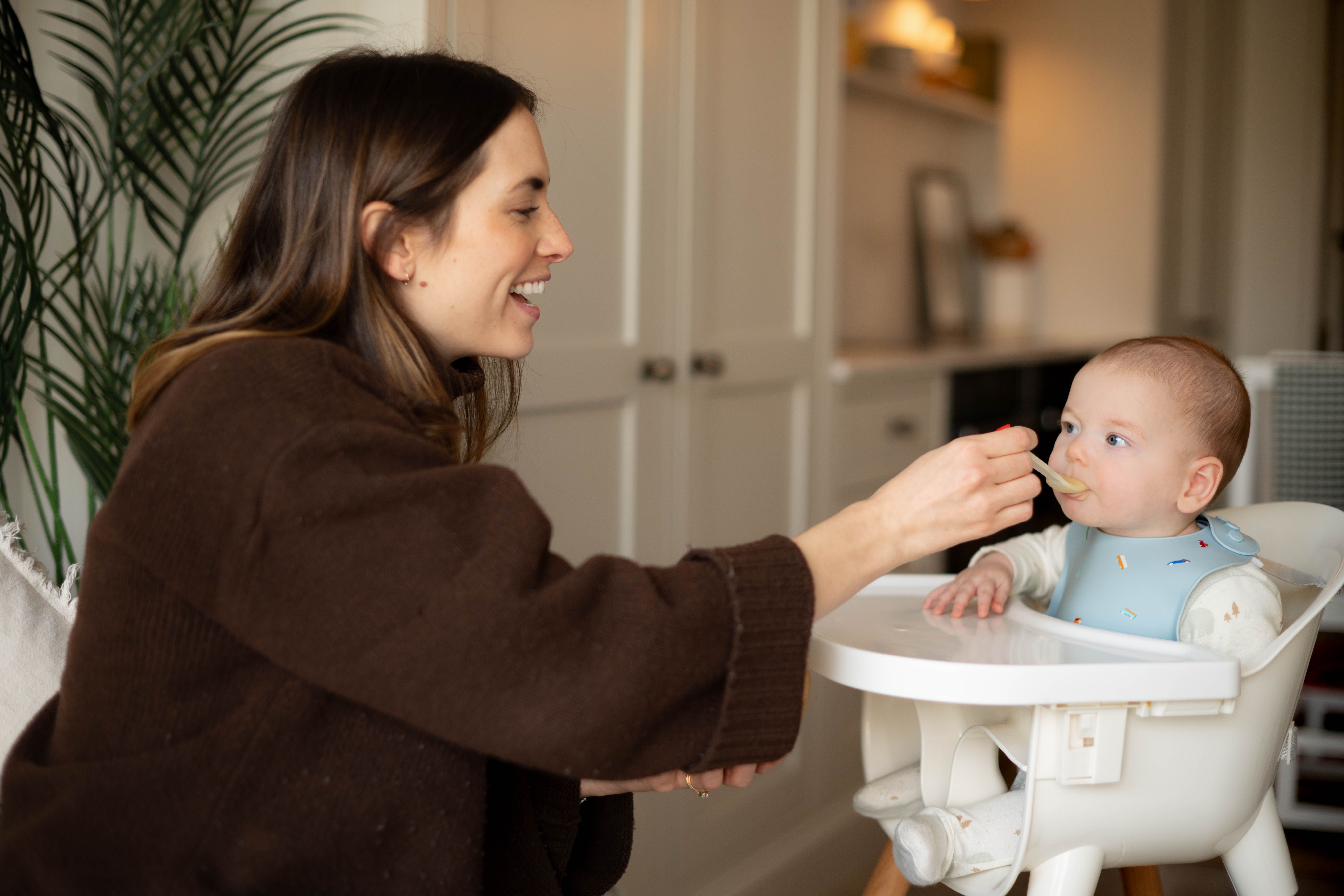 Smiling parent gently spoon-feeding first solids to baby in a highchair during early weaning.