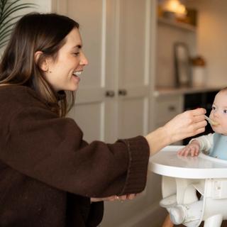 Smiling parent gently spoon-feeding first solids to baby in a highchair during early weaning.