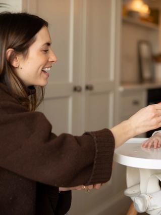 Smiling parent gently spoon-feeding first solids to baby in a highchair during early weaning.